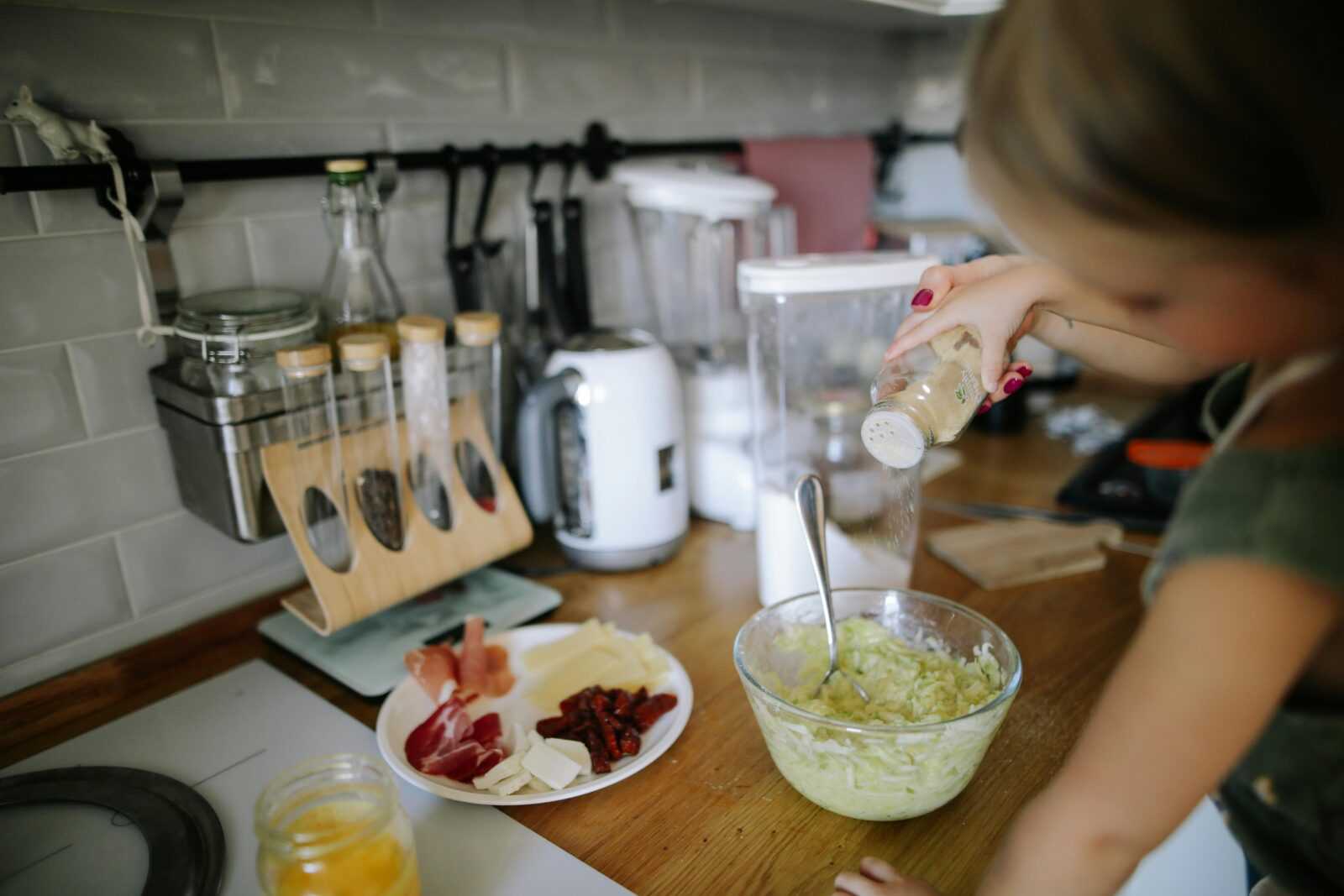 A young girl adds seasoning to a bowl in a well-equipped modern kitchen, engaging in cooking activities.