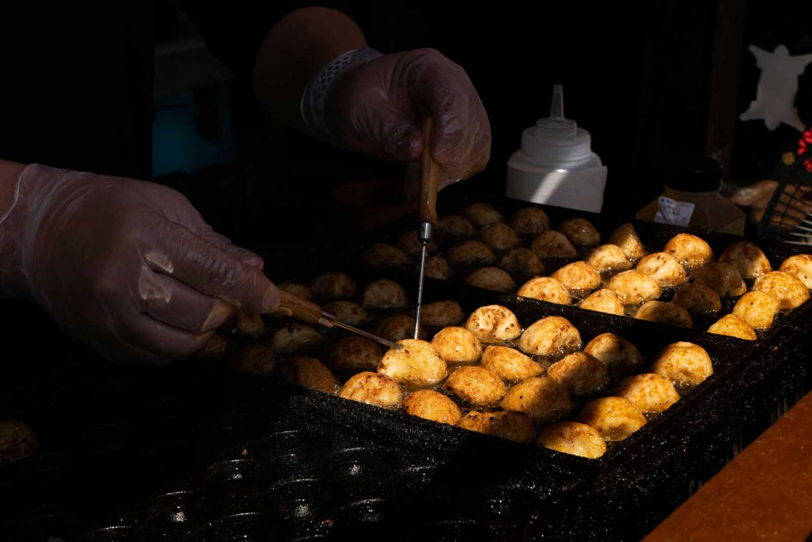 Hands turning takoyaki balls on a hot griddle at a Japanese street food stall.